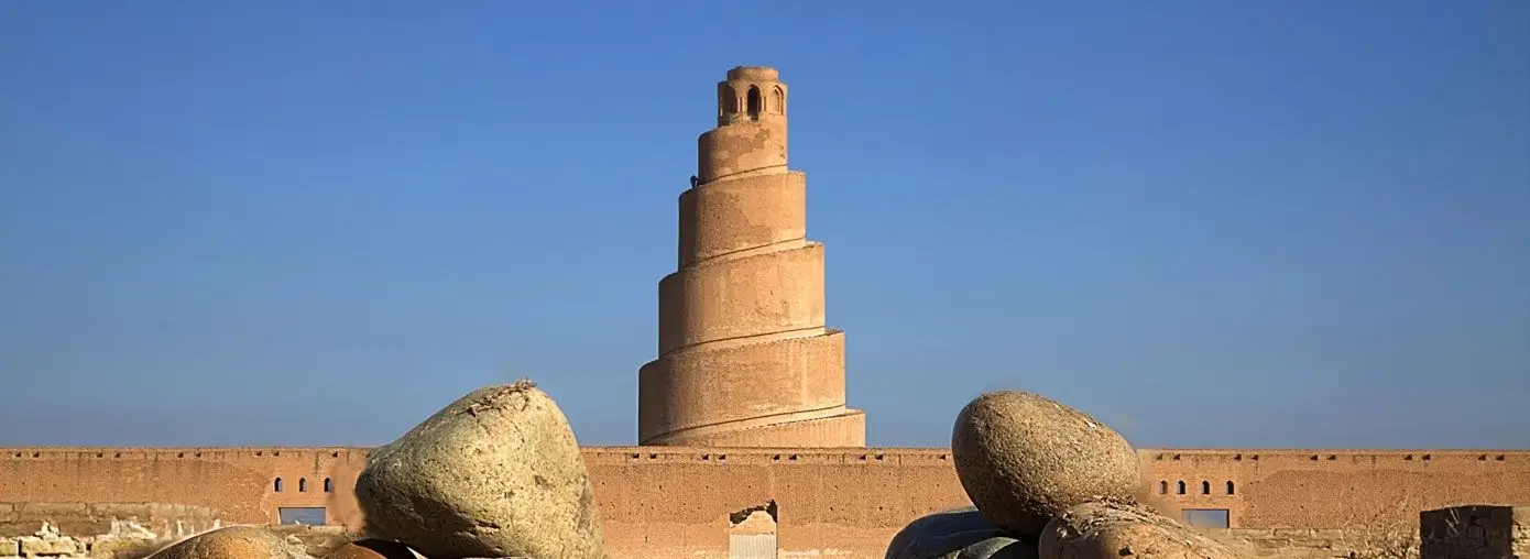 The golden dome and twin golden minarets of the Al-Askari Shrine in Samarra rising above the surrounding city under a soft daytime sky.